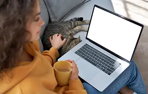 A woman sitting on a couch with a cat on her lap, using a laptop and holding a cup of coffee