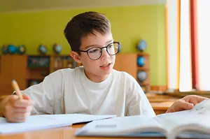 A young boy wearing glasses is sitting on a chair inside a classroom and holding a pencil and writing in a notebook while looking at a book in front of him.
