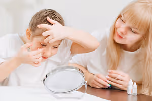 Two girls are examining their eyes in a mirror, one of them is rubbing her eye. They are sitting on a wooden table with two bottles of eye drops.
