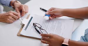 Two people, one holding a bottle of eye drops and the other holding a pair of glasses, are sitting at a table with a clipboard and paper. A watch and pen are also on the table.