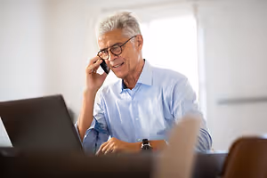 An elderly man in a blue shirt is sitting in a chair and talking on a phone