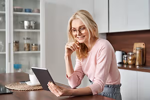 An elderly woman wearing glasses sits at a table and reads from her tablet in her kitchen