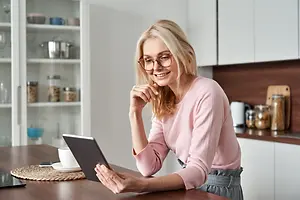 An elderly woman wearing glasses sits at a table and reads from her tablet in her kitchen