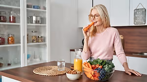 A woman eating a carrot in her kitchen. A bowl of fruits and vegetables is in front of her.