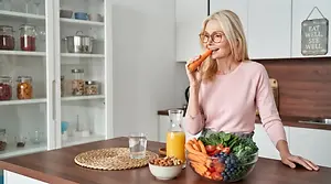 A woman eating a carrot in her kitchen. A bowl of fruits and vegetables is in front of her.