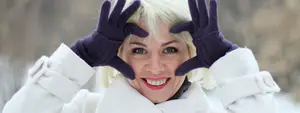 Blonde woman wearing a white coat and purple gloves smiling while posing in a snowy landscape
