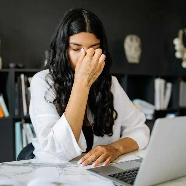 woman in white shirt sitting at desk with laptop on it with her hand covering her nose and eyes