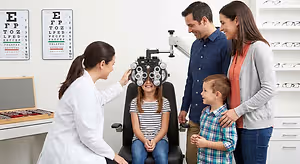 A family visiting an optometrist for a routine eye exam with the mother and daughter smiling and the father and son looking at each other while the doctor examines the daughter's eyes.
