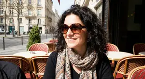 A woman with curly hair wearing a scarf and sunglasses sits at a cafe table in Paris, France.