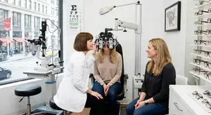 Two women and a patient are sitting in an eye exam room. One woman is testing the patient's vision with an ophthalmoscope.