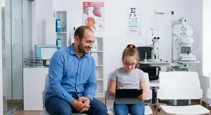 An adult man and a young girl are sitting in a medical office. The man is wearing a blue shirt and jeans. The girl is wearing glasses and a gray t-shirt. They are looking at a tablet. Behind them are medical equipment and a white shelf. The wall has an eye chart and a diagram of the human eye.