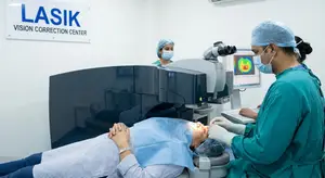 A person is lying on a table with a medical instrument over their eyes while two people wearing surgical caps and masks work on them.