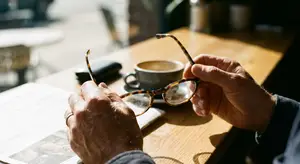 An older person holding a pair of glasses while sitting at a table and reading a newspaper.