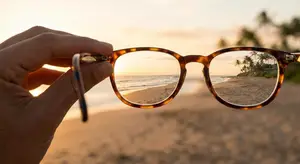 A pair of glasses reflects a beach scene, with a beach, trees, and a setting sun in the distance.