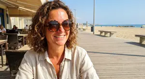 A woman with curly hair wearing sunglasses and a white shirt smiles for a selfie on a boardwalk near the beach
