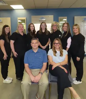Nine adults, six women and one man, are smiling in front of a blue wall and a mirror.