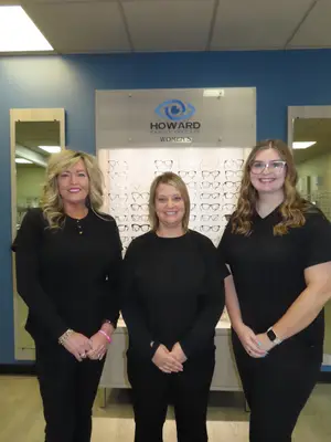 Three women are standing in front of a display of women's eyeglasses in an office setting.