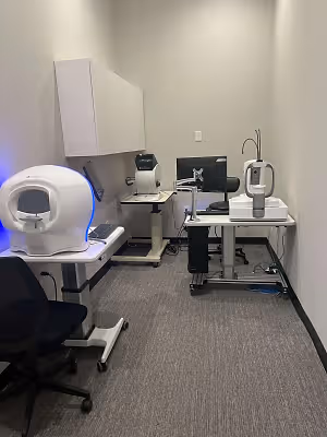 A medical examination room equipped with an advanced diagnostic machine, a computer monitor, and various medical devices, set against a white wall and gray carpet.