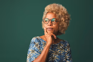 A woman with curly blonde hair wearing glasses and a blue tropical shirt is posing for a photo in front of a green background.