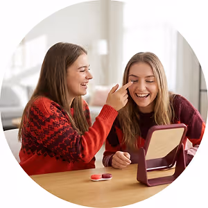 Two women sitting at a table with a small mirror, smiling and looking at each other with one putting a contact lens in the other's eye