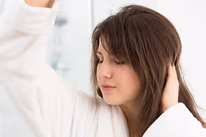 A woman in a white robe is closing her eyes in a bathroom while holding her hair with her right hand.