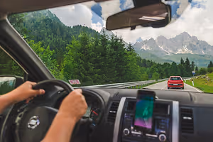 Driver driving on mountain road with mountains and lush greenery in the background, a red car in front, and a dashboard view with a cell phone, steering wheel, and speedometer.