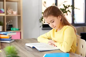 A girl is sitting on a chair while reading a book on a desk.
