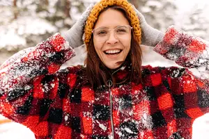 A smiling woman wearing a red and black plaid coat and a yellow hat covered in snow stands in the snow.