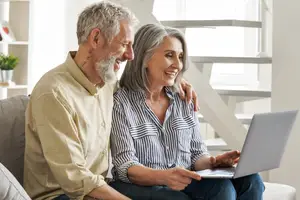 An older couple sits on a couch, with the woman using a laptop and the man looking at her with a smile.