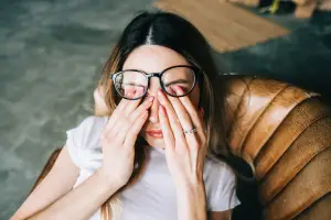 A woman sitting on a brown leather couch, wearing glasses, covering her eyes with her hands, and looking sideways