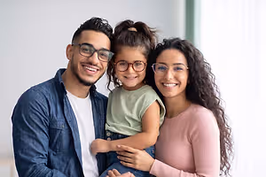 A man and a woman with a young girl in between them, all wearing eyeglasses, standing close to each other with a smile, probably in the room