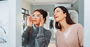 Two women wearing glasses are standing in a room and looking at something. One of them is holding her face with both hands, and the other is holding her glasses with her left hand. Behind them is a glass door with a reflection of a person.