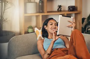 A woman is smiling and holding a tablet in her hands while sitting on a couch and seems to be posing for a photo.