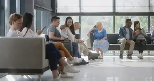 People sitting on chairs and benches in a waiting area, some are holding cell phones and others are looking at them.