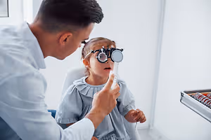 A man is examining a young girl's eye with an ophthalmoscope in a clinic.