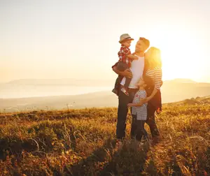 a man holding a toddler and a girl standing next to him with a woman behind them in a grassy field with a beautiful sunset in the background