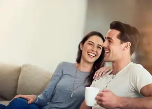 A man and woman are sitting on a couch, smiling and holding a white cup, possibly in a living room