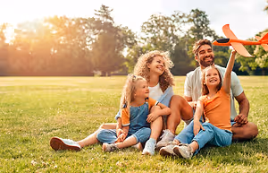 A family of four is sitting on the grass in a park, with a man holding a toy airplane, and the two girls are smiling and posing for a photo.