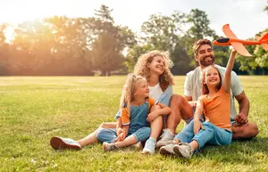 A family of four is sitting on the grass in a park, with a man holding a toy airplane, and the two girls are smiling and posing for a photo.