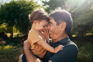 A man wearing a black polo shirt is holding a child wearing a brown shirt while both of them are smiling in a grassy area with trees and plants in the background.