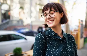 Woman with glasses and a polka dot shirt smiles on the sidewalk