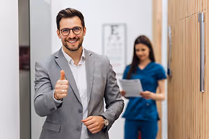 A man in a gray suit gives a thumbs up in a medical office, while a woman in blue scrubs stands in the background.