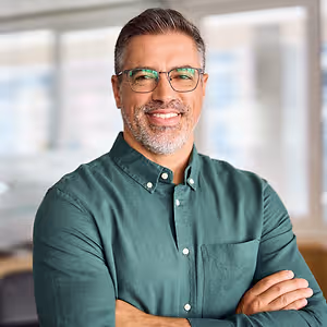 A man wearing glasses and a green polo shirt with his arms crossed and smiling while standing in an office.