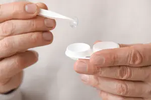 A man's hands hold a lens in a container and a dropper with a liquid inside it.