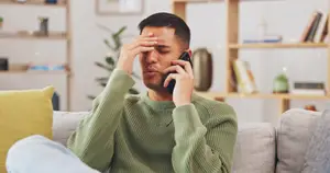 A man on a couch is holding a phone to his ear and his other hand is covering his forehead. He is looking at something on the phone. He is wearing a green sweater. Behind him is a shelf with books and some other objects. There is also a potted plant in the corner of the room. The room has a white wall.