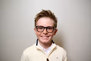 A smiling young boy with glasses and a light beige jacket is posing for a photo against a white background.