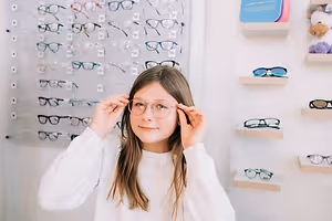 A young girl trying on glasses in an optical shop with a display of different frames behind her.