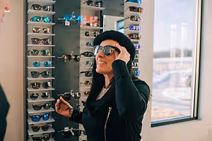 A woman in a black outfit is standing in front of a wall of sunglasses, smiling, and adjusting her sunglasses.