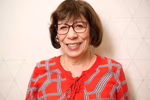 Portrait of a smiling woman in a red patterned shirt and glasses against a white geometric wall background