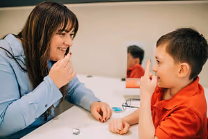 A woman and a young boy are seated at a table, with the woman showing something to the boy and both of them smiling, and a mirror reflecting another person behind them.